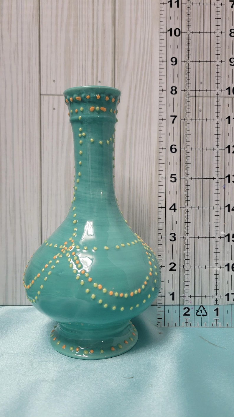 K&ouml;nnte beinhalten: Eine t&uuml;rkisfarbene Keramikvase mit bauchigem Boden und schlankem Hals. Die Vase ist mit kleinen, erhabenen orangefarbenen und gelben Punkten verziert. Die Vase ist etwa 20 cm hoch.