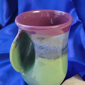 Puede incluir: Una taza de cerámica hecha a mano con un diseño de asa único. La taza presenta un degradado de color, que va desde una base verde claro hasta un centro azul-gris, y un borde morado oscuro. La taza mide aproximadamente 10 cm de alto.