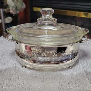 Peut inclure: Un plat à gratin ovale en verre avec un couvercle en verre transparent doté d'un bouton. Le plat est inséré dans une monture en métal argenté avec des détails en filigrane découpés, deux anses latérales et quatre petits pieds.
