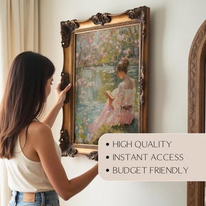 May include: A framed painting with a gold ornate frame is being hung on a wall. The painting depicts a woman reading near a body of water with pink blossoms. The frame is a dark gold color.