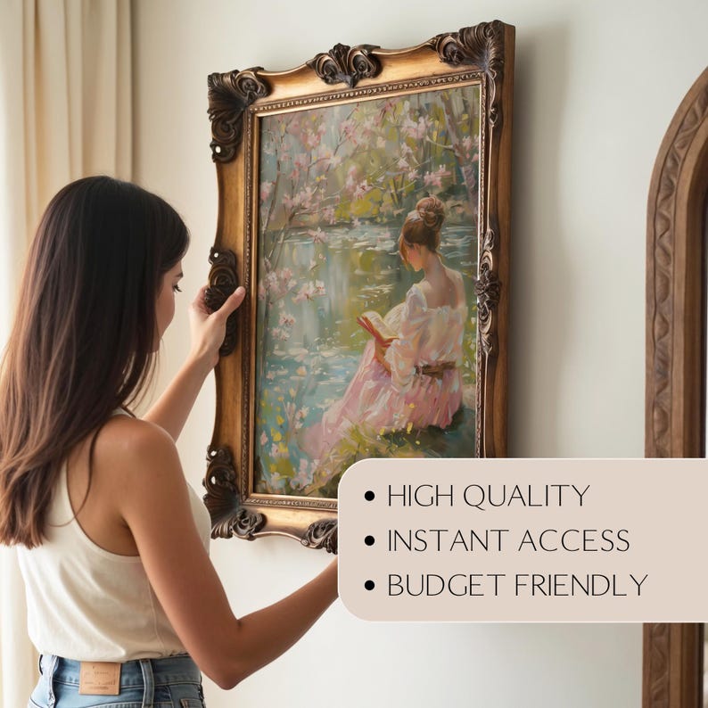 May include: A framed painting with a gold ornate frame is being hung on a wall. The painting depicts a woman reading near a body of water with pink blossoms. The frame is a dark gold color.