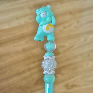 May include: A decorative pen featuring a light teal bear figurine topper with a yellow rainbow emblem on its chest. The pen barrel is adorned with a series of beads, including a clear sphere, a white textured floral bead, and a teal heart bead.