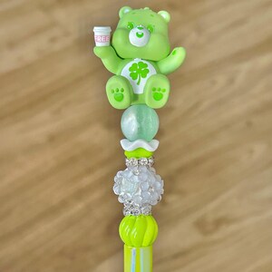 May include: A lime green bear-shaped charm with a four-leaf clover on its belly, holding a small white cup with the word "FREE" printed on it. The charm is attached to a decorative stick with various beads in white, green, and silver.