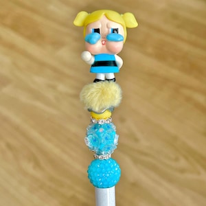 May include: A decorative pen featuring a cartoon character figurine with yellow hair and a blue and black outfit. The pen has a yellow pom-pom, blue and clear beads, and a silver-colored base. The pen is designed for writing.