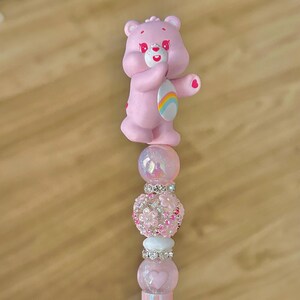 May include: A pink Care Bear figurine tops a beaded accessory. The accessory features iridescent and floral beads, along with a heart-shaped bead. The Care Bear has a rainbow on its belly.