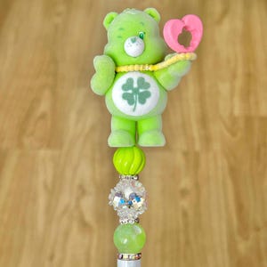 May include: A green Care Bear-themed pen with a four-leaf clover on its belly and a pink heart. The pen features a series of decorative beads in shades of green and white, with a silver-colored base.