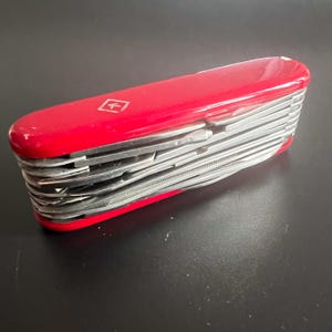 Könnte beinhalten: Ein rotes Schweizer Taschenmesser mit mehreren sichtbaren Werkzeugen. Das Messer hat eine rechteckige Form mit abgerundeten Kanten und einem kleinen Emblem oben. Die Werkzeuge sind aus silberfarbenem Metall und teilweise ausgeklappt.