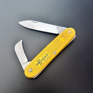 以下が含まれることがあります： 2つの刃が開いた黄色のポケットナイフ。ナイフは「Bugnard」の文字と十字のロゴが入ったテクスチャード加工のハンドルが付いています。刃は銀色で、ナイフは黒い表面にあります。