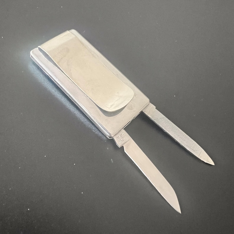 May include: A silver pocket knife with a money clip. The knife features a blade and a nail file. The metal knife has a shiny finish.