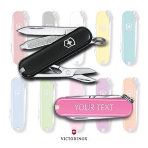 Può includere: Un coltellino tascabile nero e argento con un logo a croce rossa e un piccolo paio di forbici. Il coltellino è aperto e la lama è visibile. Il coltellino è circondato da una varietà di coltellini tascabili colorati, tra cui rosa, blu, verde e giallo. Il testo "YOUR TEXT" è inciso sul coltellino tascabile rosa.