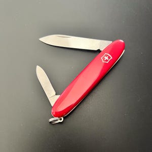 Puede incluir: Una navaja suiza roja con dos hojas abiertas y una hoja cerrada. La navaja tiene una hoja plateada y un mango rojo con un emblema de cruz blanca. La navaja está sobre una superficie oscura.