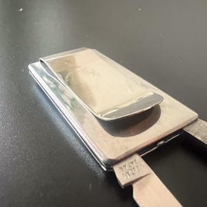 May include: A silver money clip with a hinged design. The clip has a rectangular base with a curved top, and a smaller, hinged section. The text "VULCAN JAPAN" is stamped on the side.