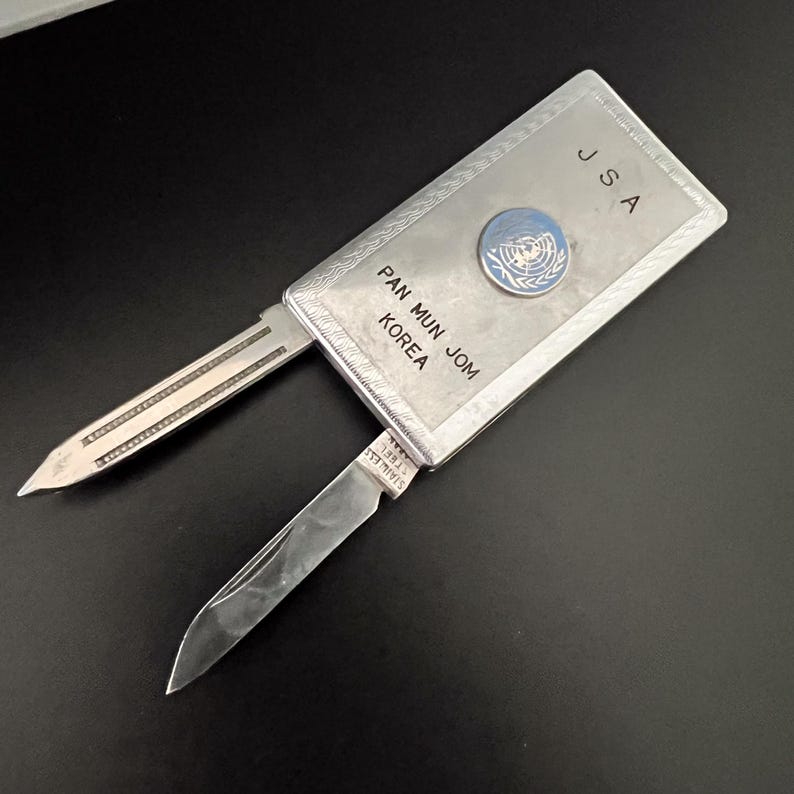 May include: A silver pocket knife with two blades extended. The knife has the text "JSA" and "PAN MUN JOM KOREA" engraved on the handle, along with a United Nations emblem. The blades are stainless steel.