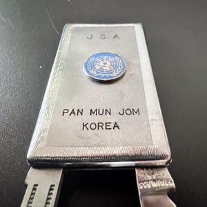 May include: A silver-coloured metal object with the text "JSA" and "PAN MUN JOM KOREA". A blue and white United Nations emblem is centered on the top. The bottom portion has a stainless steel blade.
