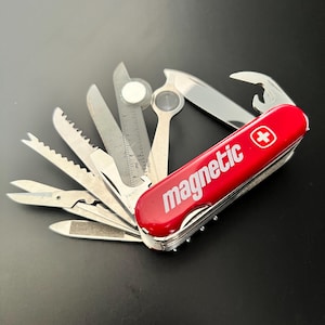 May include: A red multi-tool pocket knife with the word magnetic and a white cross logo printed on the side. The tool is fully opened, displaying various stainless steel implements including a saw, a ruler, a magnifying glass, and a blade.