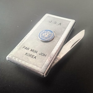 Peut inclure: Un couteau de poche argenté avec les inscriptions "JSA" et "PAN MUN JOM KOREA" gravées sur le devant. Un emblème bleu et blanc des Nations Unies est apposé sur le devant. La lame du couteau est partiellement ouverte.