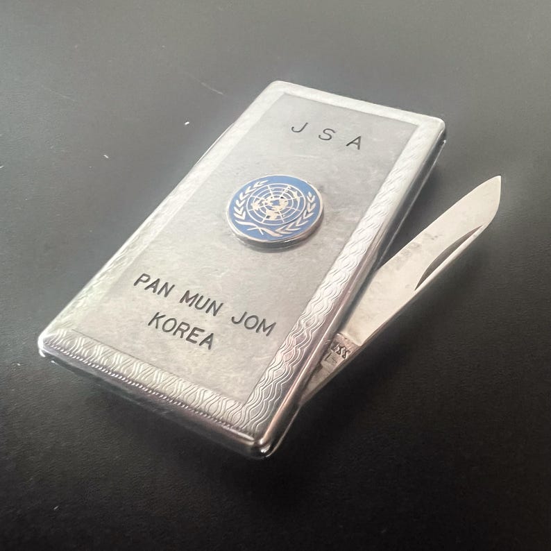 May include: A silver pocket knife with the text "JSA" and "PAN MUN JOM KOREA" engraved on the front. A blue and white United Nations emblem is affixed to the front. The knife blade is partially open.
