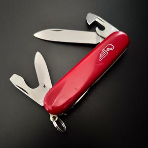 Könnte beinhalten: Ein rotes Multitool-Messer mit mehreren Edelstahlklingen und Werkzeugen. Das Messer enthält eine Klinge, einen Flaschenöffner und andere Werkzeuge. Der rote Griff hat ein weißes Logo und einen Schlüsselring.