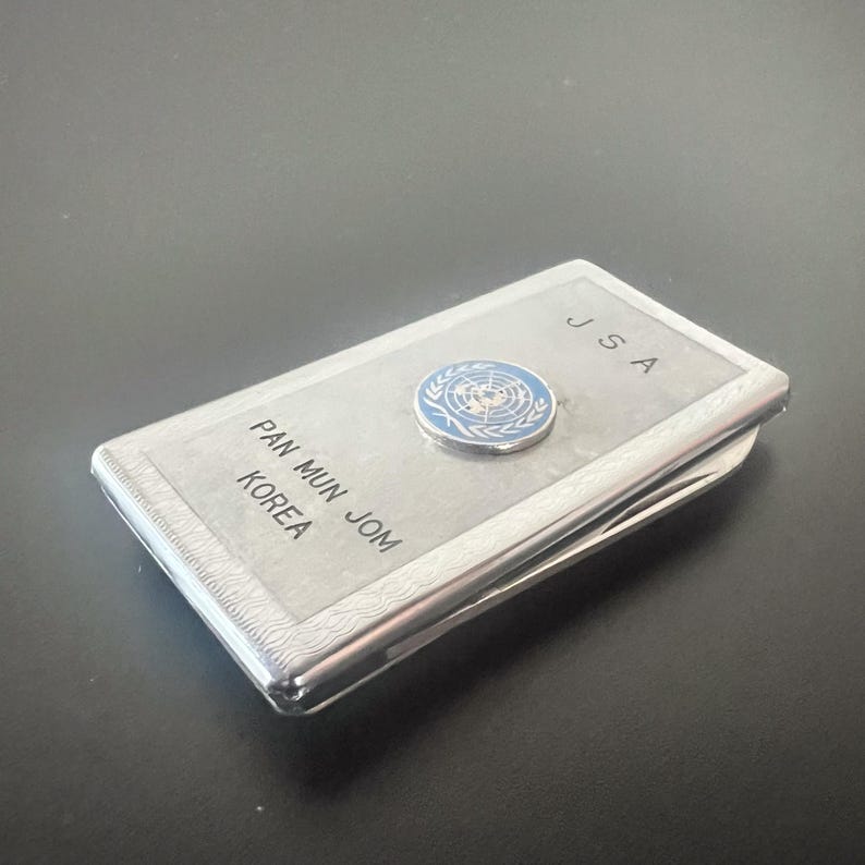 May include: A silver cigarette case with the text "JSA" and "PAN MUN JOM KOREA" engraved on the front. A blue and white United Nations emblem is affixed to the top. The case is rectangular and has a polished finish.