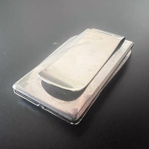 May include: A silver money clip, featuring a rectangular base and a curved, spring-loaded clip. The clip is made of polished metal, designed to securely hold folded banknotes. The design is simple and elegant, suitable for everyday use.