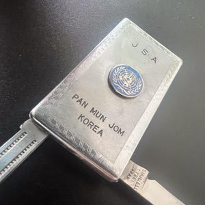 May include: A silver multi-tool with the text "JSA" and "PAN MUN JOM KOREA". It features a blue and white emblem and a stainless steel blade. The tool is rectangular with decorative edges.