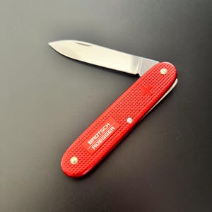 以下が含まれることがあります： 銀色の刃が付いた赤いポケットナイフ。テクスチャード加工されたハンドルと「BROTSCH ROEGGER」の文字が特徴です。ナイフには小さな赤い十字のエンブレムがあり、開いていて刃が見えています。