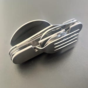 May include: A compact, multi-tool utensil set in a matte silver finish. The set includes a fork, spoon, knife, and a bottle opener, all neatly nested together. The utensils are made of stainless steel and are designed for portability and convenience.