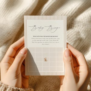 May include: A rectangular Baby Bingo game card with a beige and white striped border. The card features the text Baby Bingo, instructions for the game, and a five-by-five grid with a small brown teddy bear illustration in the center square.