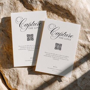 Puede incluir: Dos tarjetas de boda de color marfil con el texto "Capture the Love" en escritura elegante. Cada tarjeta presenta un código QR y texto adicional. Las tarjetas están colocadas sobre una superficie texturizada de color claro.