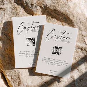 May include: Two off-white cards with the text "Capture the Love" in elegant script. Each card features a QR code and a message inviting guests to share photos. The cards are placed on a textured, light-colored surface.