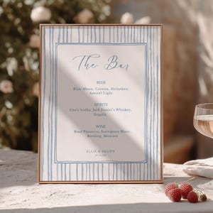 May include: A rectangular bar menu sign in a thin gold frame featuring a light beige background with blue vertical stripes. The text reads The Bar, Beer, Spirits, Wine, and the names Ellie & Shawn with the date 07.16.XX in a blue script and serif font.