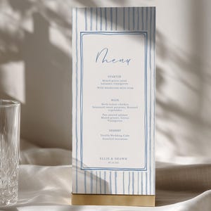 May include: A vertical rectangular menu card featuring a light blue and white striped border with a hand-drawn blue frame. The text reads Menu, Starter, Main, Dessert, and Ellie & Shawn 07.12.24. The card is held upright by a gold-colored metal base.