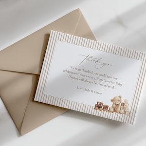 May include: A beige envelope and a rectangular thank you card featuring a light brown striped border. The card displays the text Thank you, We're so thankful you could join our celebration! Your sweet gift and love for Baby [Name] will always be remembered. Love, Jade & Chase, with a small illustration of a teddy bear, a toy train, and alphabet blocks.