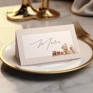 May include: A folded place card with a beige and white vertical striped border. The center features the name Maria in black cursive script, accompanied by a small watercolor illustration of a brown teddy bear, a toy train, and alphabet blocks.