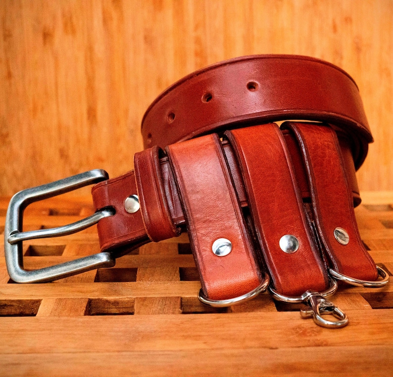 May include: A brown leather belt with multiple straps and a silver buckle. The belt has three D-rings and a silver hook.
