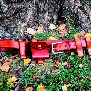 May include: A brown leather belt with a small brown leather pouch attached. The belt is lying on a bed of green grass and brown leaves.