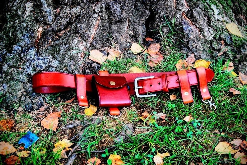 May include: A brown leather belt with a small brown leather pouch attached. The belt is lying on a bed of green grass and brown leaves.