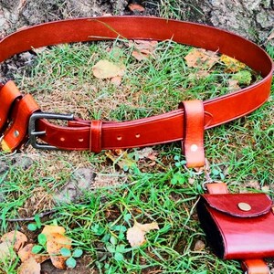 May include: A brown leather belt with a silver buckle and multiple leather pouches attached. The belt is lying on a bed of green grass and brown leaves.