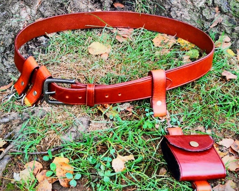 May include: A brown leather belt with a silver buckle and multiple leather pouches attached. The belt is lying on a bed of green grass and brown leaves.