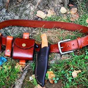 May include: A brown leather belt with a silver buckle and two leather pouches. A leather knife sheath with a wooden-handled knife is attached to the belt.