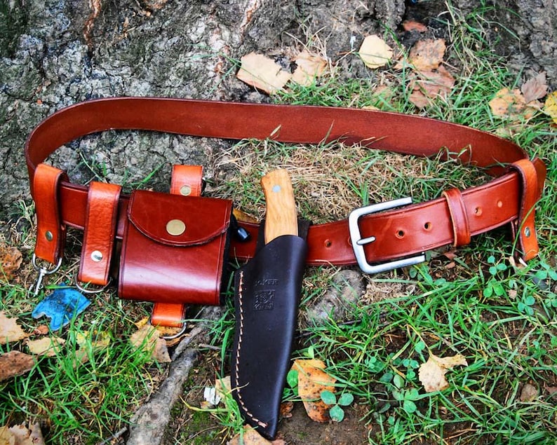 May include: A brown leather belt with a silver buckle and two leather pouches. A leather knife sheath with a wooden-handled knife is attached to the belt.