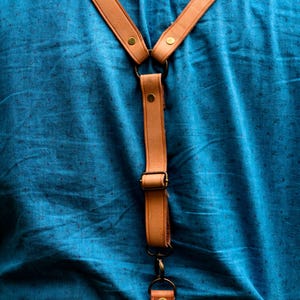 Könnte beinhalten: Ein braunes Lederkameraband mit verstellbaren Schnallen und einem Metallclip. Das Band ist an einem blauen Hemd befestigt.