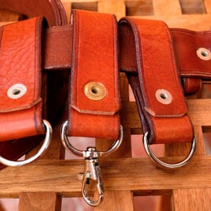 May include: Four brown leather belt loops with silver metal rings and a silver metal clip. The loops are attached to a brown leather belt.