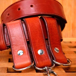 May include: Three brown leather belts with silver buckles and metal loops. The belts are stacked on top of each other.
