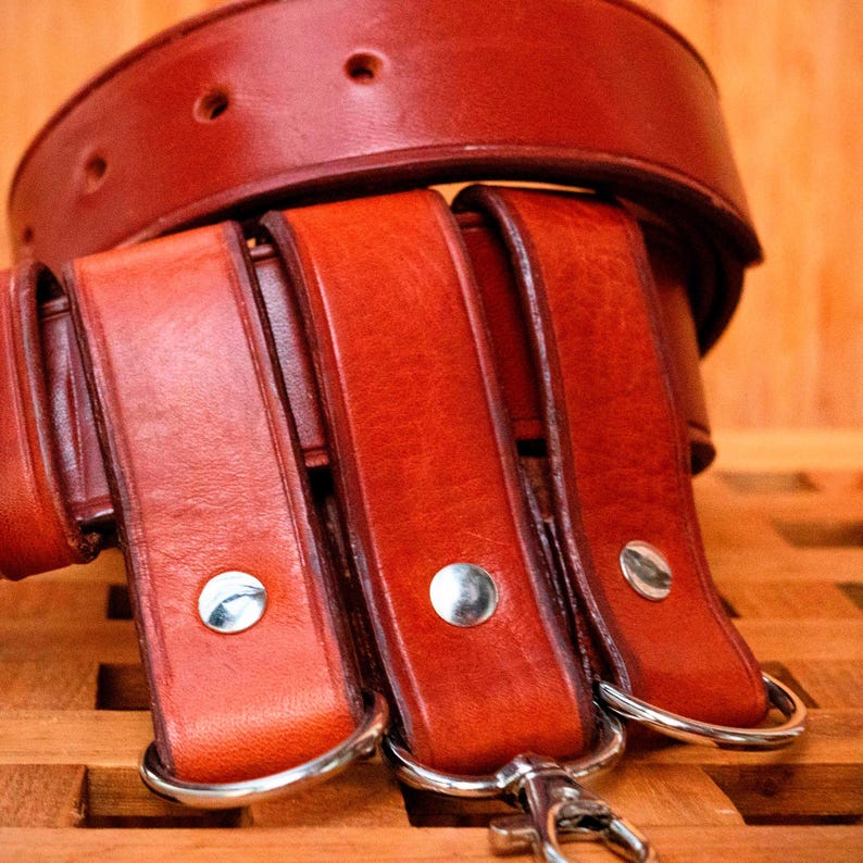 May include: Three brown leather belts with silver buckles and metal loops. The belts are stacked on top of each other.