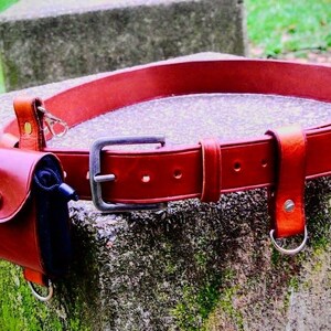May include: A red leather belt with multiple loops and a small pouch attached. The belt has a silver buckle and is decorated with stitching.