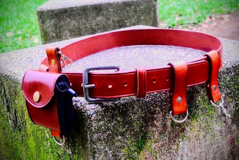 May include: A red leather belt with multiple loops and a small pouch attached. The belt has a silver buckle and is decorated with stitching.
