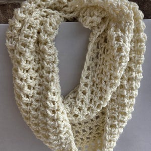 May include: Cream-colored, handmade crochet infinity scarf. The scarf is made of a thick, textured yarn, with a repeating open stitch pattern. The scarf is displayed against a neutral background.