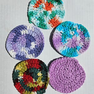May include: Five handmade, circular crochet coasters in various colors. The coasters feature a spiral design, with colors including orange, green, blue, purple, and red. The coasters are made of a soft, textured yarn.