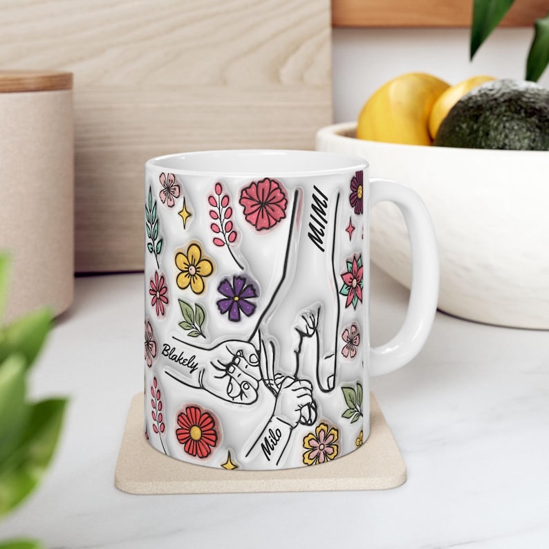 May include: White ceramic mug featuring a floral design and handprints. The mug is inscribed with the names "Mimi", "Blakely", and "Milo".